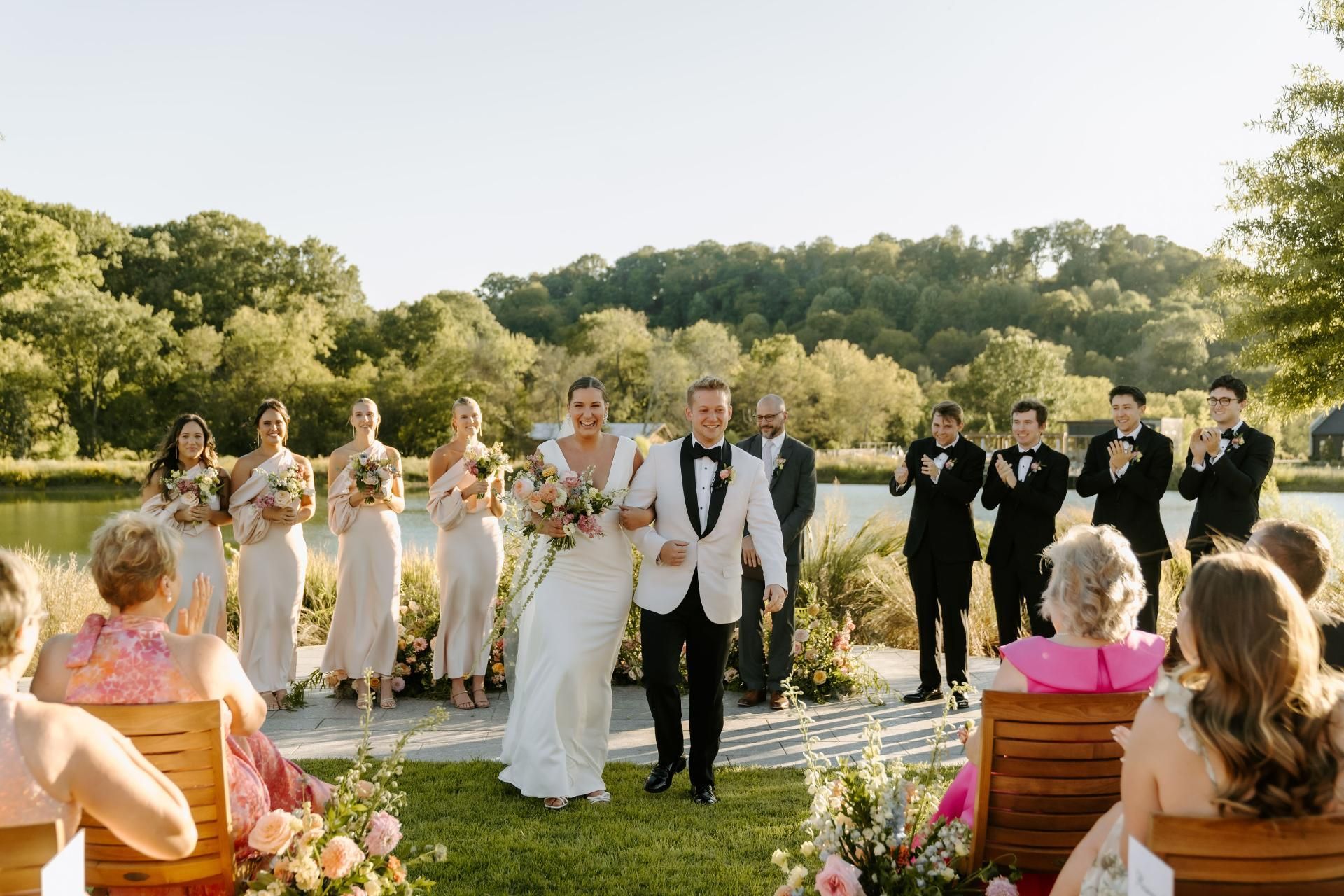 Newlyweds walk down the aisle, smiling, amidst a wedding ceremony with bridesmaids and groomsmen, lake setting.