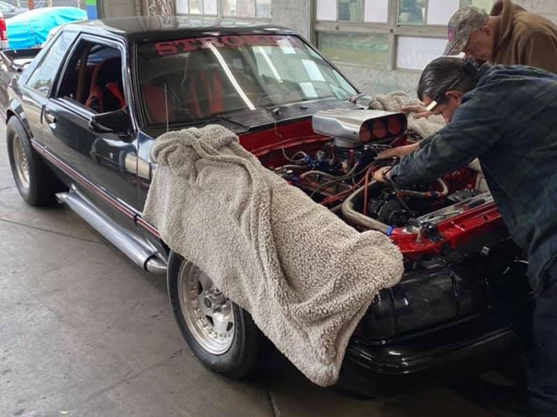 Classic black car with its hood open while two people work on the engine in a garage.