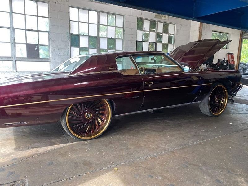 Classic dark maroon lowrider car with hood open in a garage, featuring gold-spoke wheels.