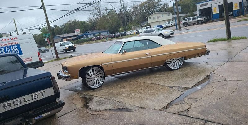 Tan vintage lowrider car on a wet street, angled on raised suspension near a parking lot.
