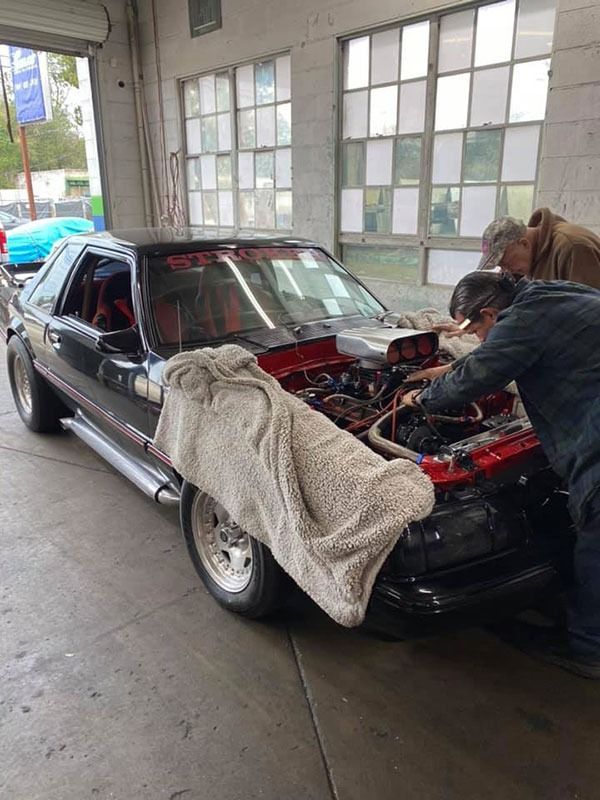 Black classic car in a garage with hood open, covered by a blanket while someone works on the engine.