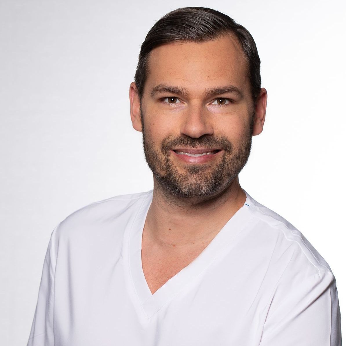 En man med mörkt hår och skägg ler, klädd i en vit V-ringad topp, mot en vit bakgrund.
