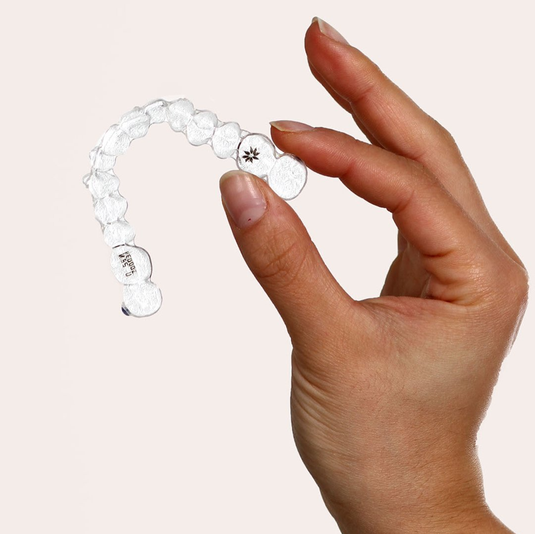 Hand som håller en genomskinlig tandriktare av plast, möjligen för tänderutjämning, mot en ljus bakgrund.