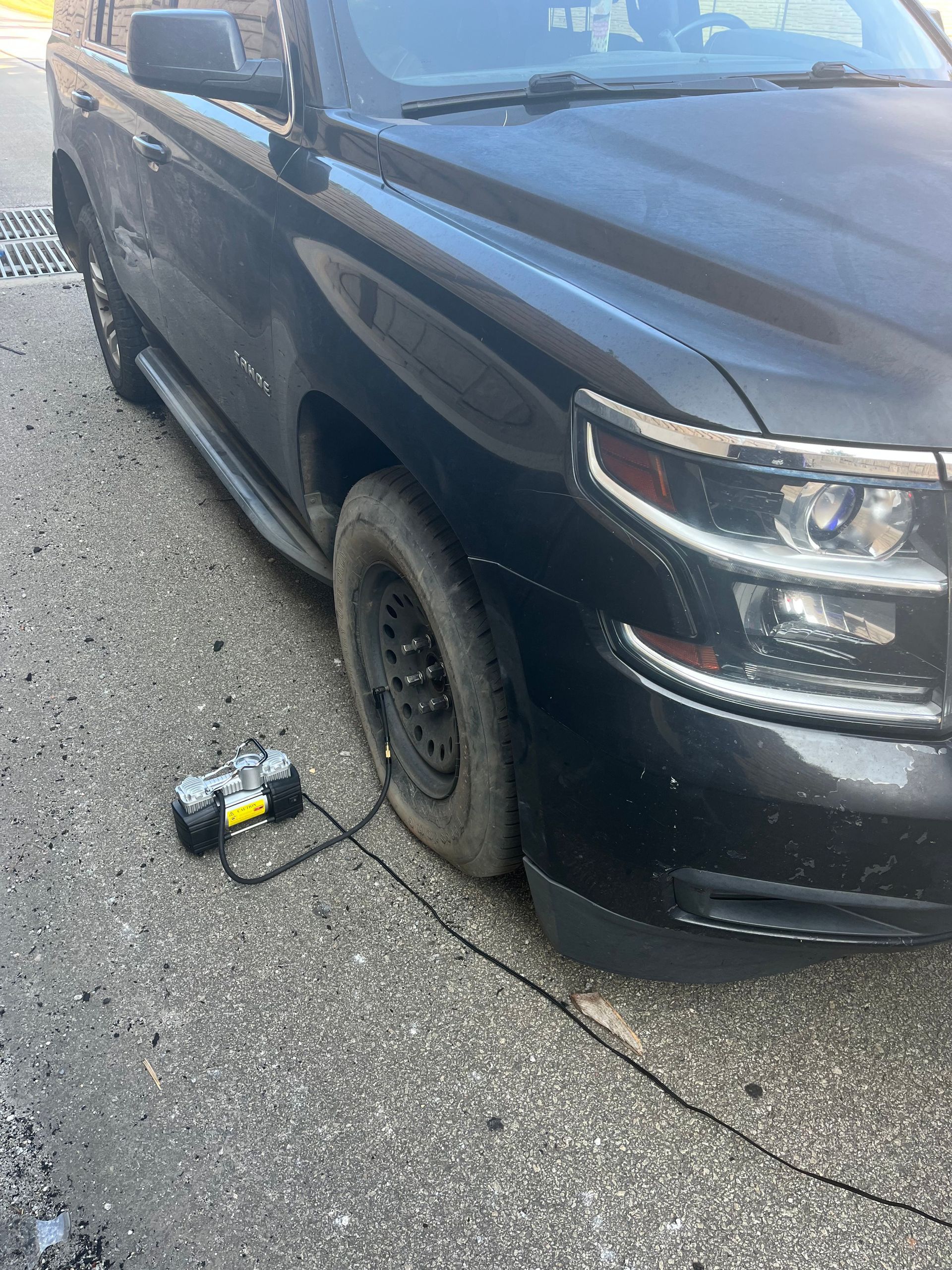 Black SUV with a flat front tire, parked on pavement beside a small portable air compressor.