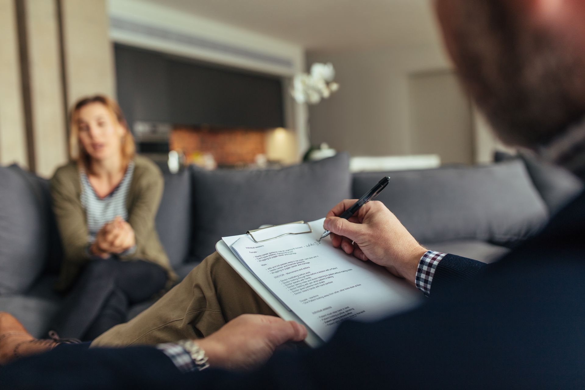 A man is writing on a clipboard while a woman sits on a couch.