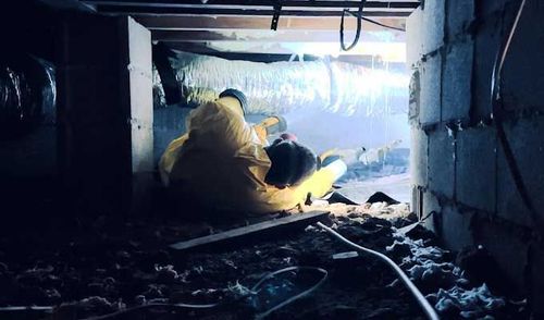 Two workers in yellow protective suits crawl in a crawl space, near insulation and concrete blocks.