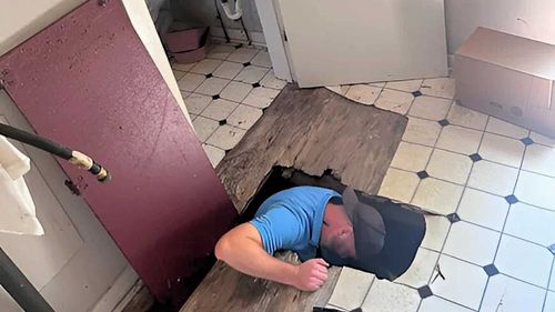Man in blue shirt and hat reaching into hole in floor of room with checkered tiles and debris.