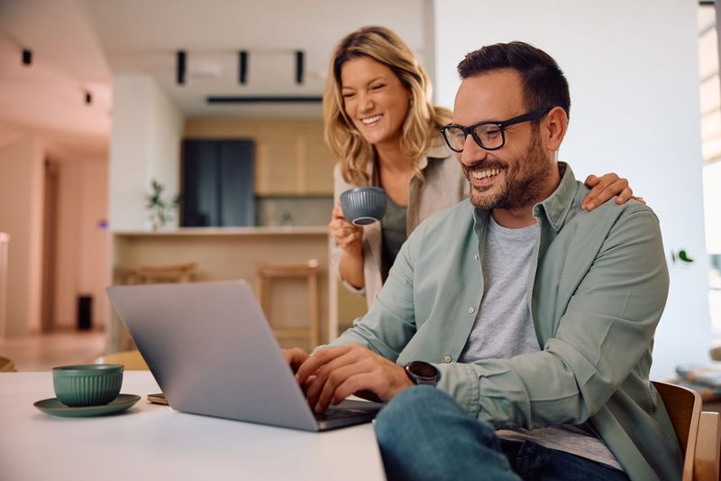 Two people smiling at a laptop in a bright kitchen, one holding a mug