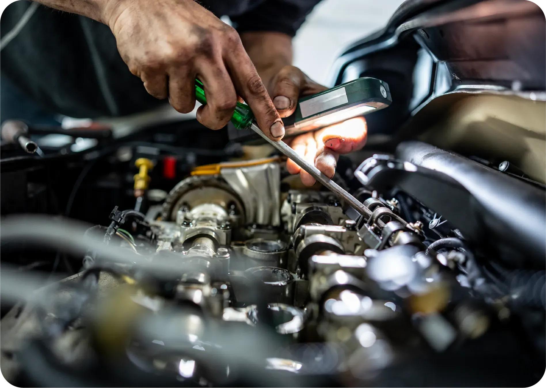 A mechanic uses a screwdriver and a small handheld light to work on the internal components of a car engine.