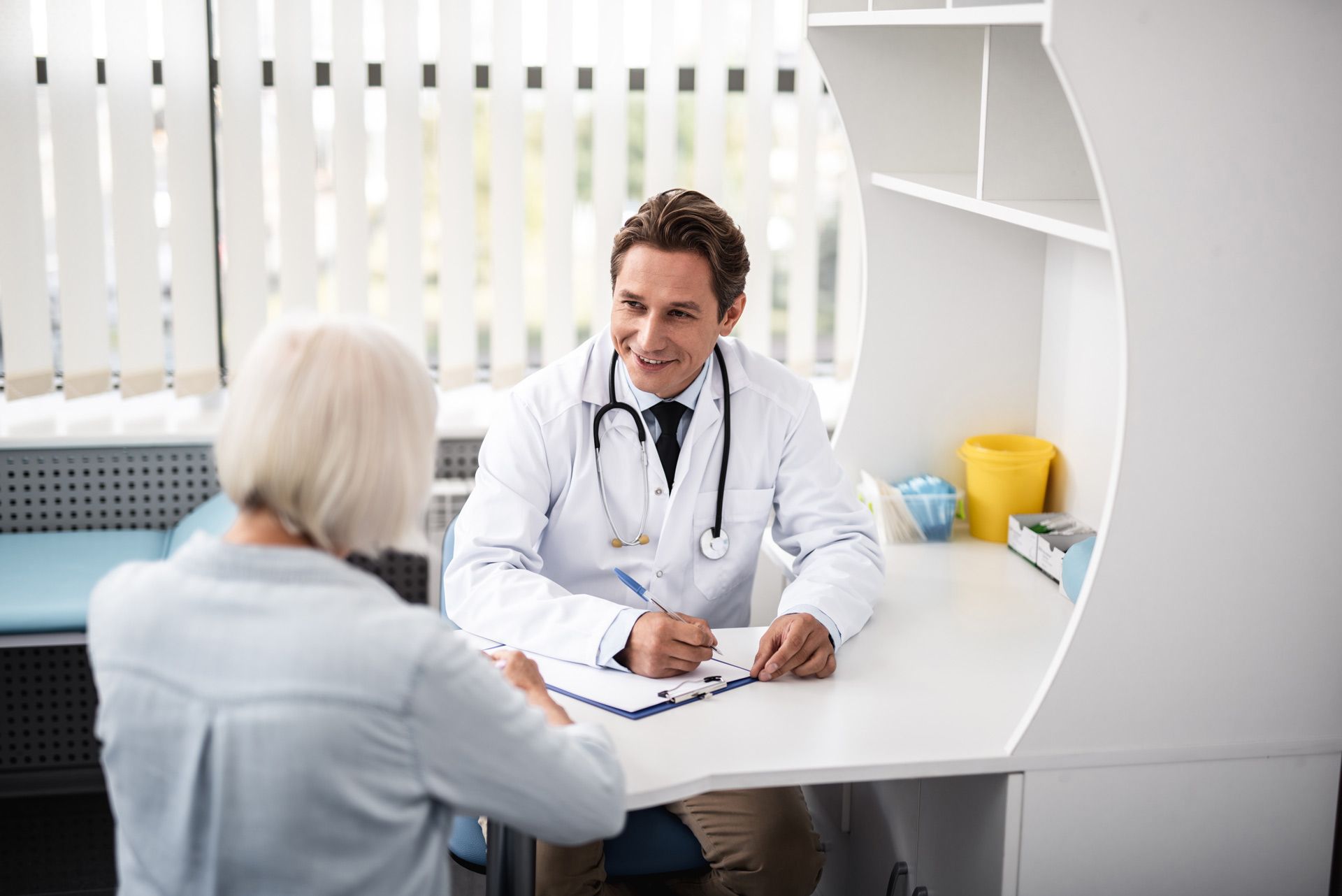 A cheerful experienced general practitioner sitting at the table with his patient, looking at her.