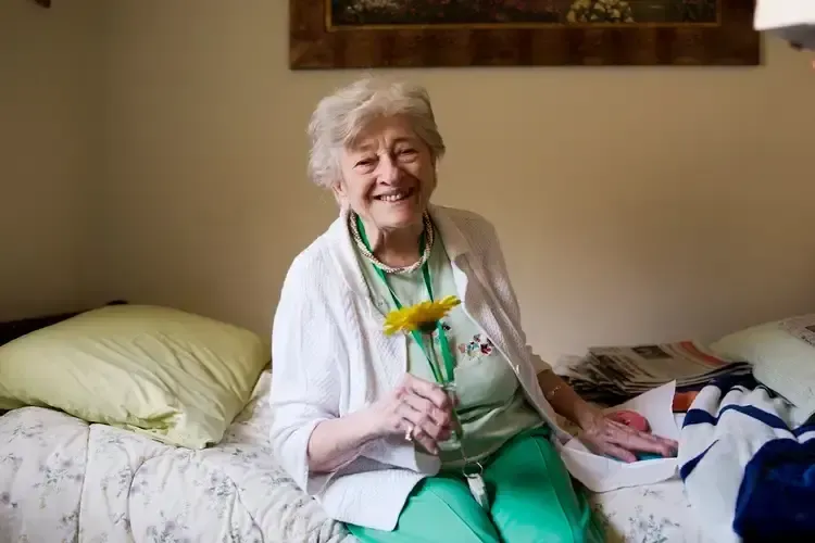 An elderly woman is sitting on a bed holding a yellow flower.