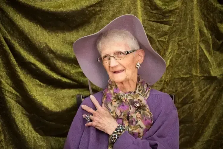An elderly woman wearing a purple hat and scarf is standing in front of a green curtain.