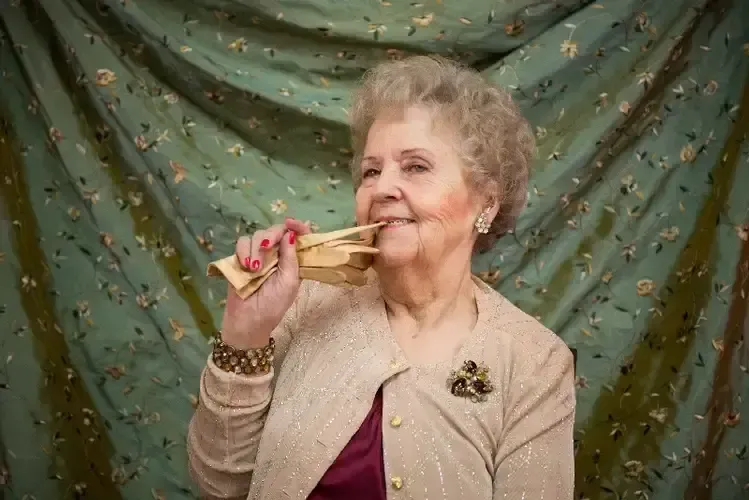 An elderly woman is blowing a party horn in front of a green curtain.