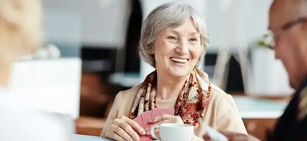 An elderly woman is sitting at a table with a cup of coffee and playing cards.