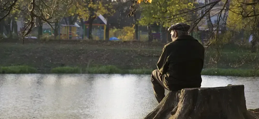 A man is sitting on a tree stump by a lake.