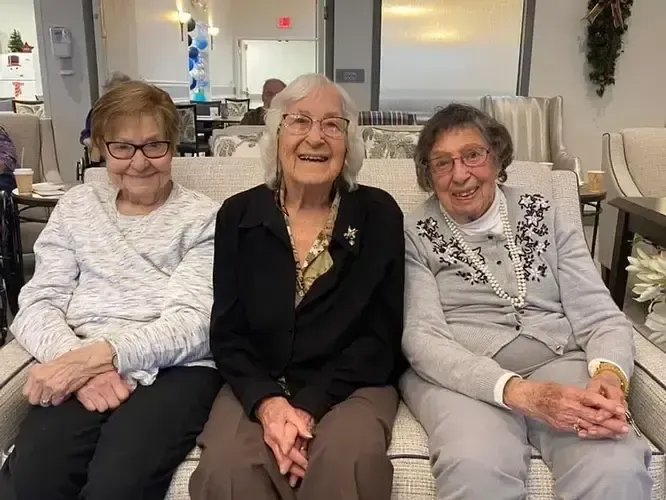 Three elderly women are sitting on a couch and smiling for the camera.