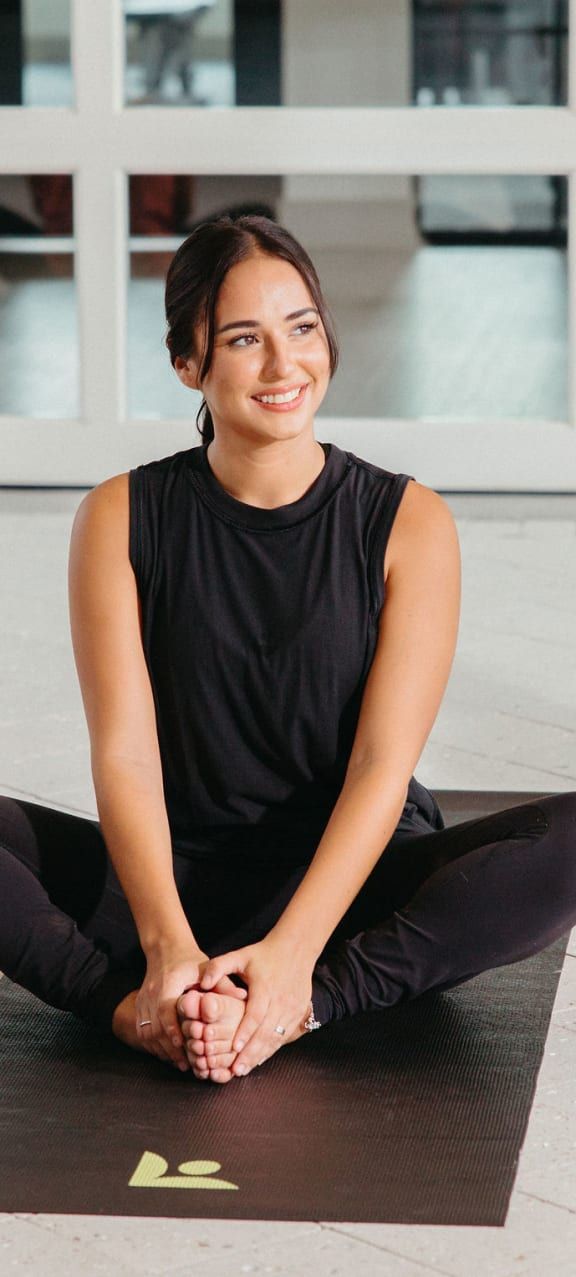 A woman is sitting on a yoga mat with her legs crossed and smiling.