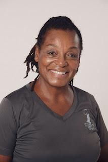 Woman with dreadlocks, smiling, wearing a gray v-neck shirt, against a white background.