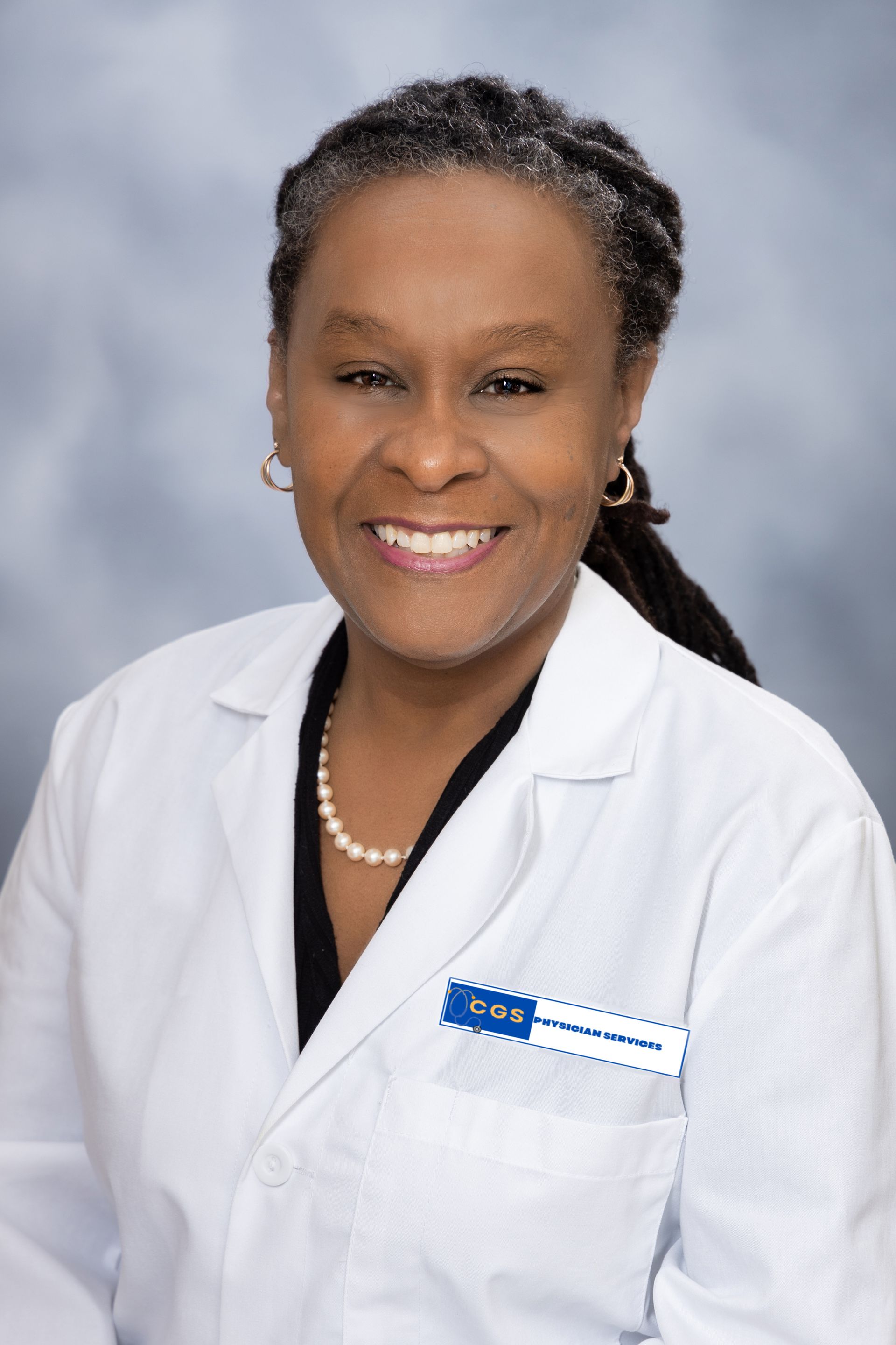 Woman in a white lab coat smiles. She wears a pearl necklace and has a name tag. Background is light blue.