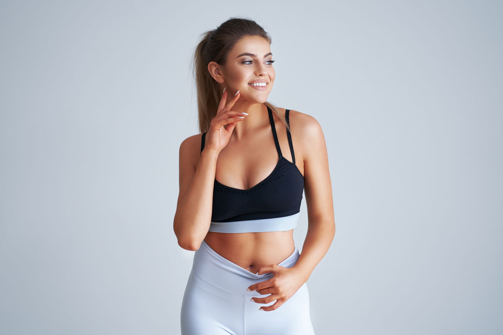 Woman in sports bra and leggings smiles, touching her face, in front of a white background.