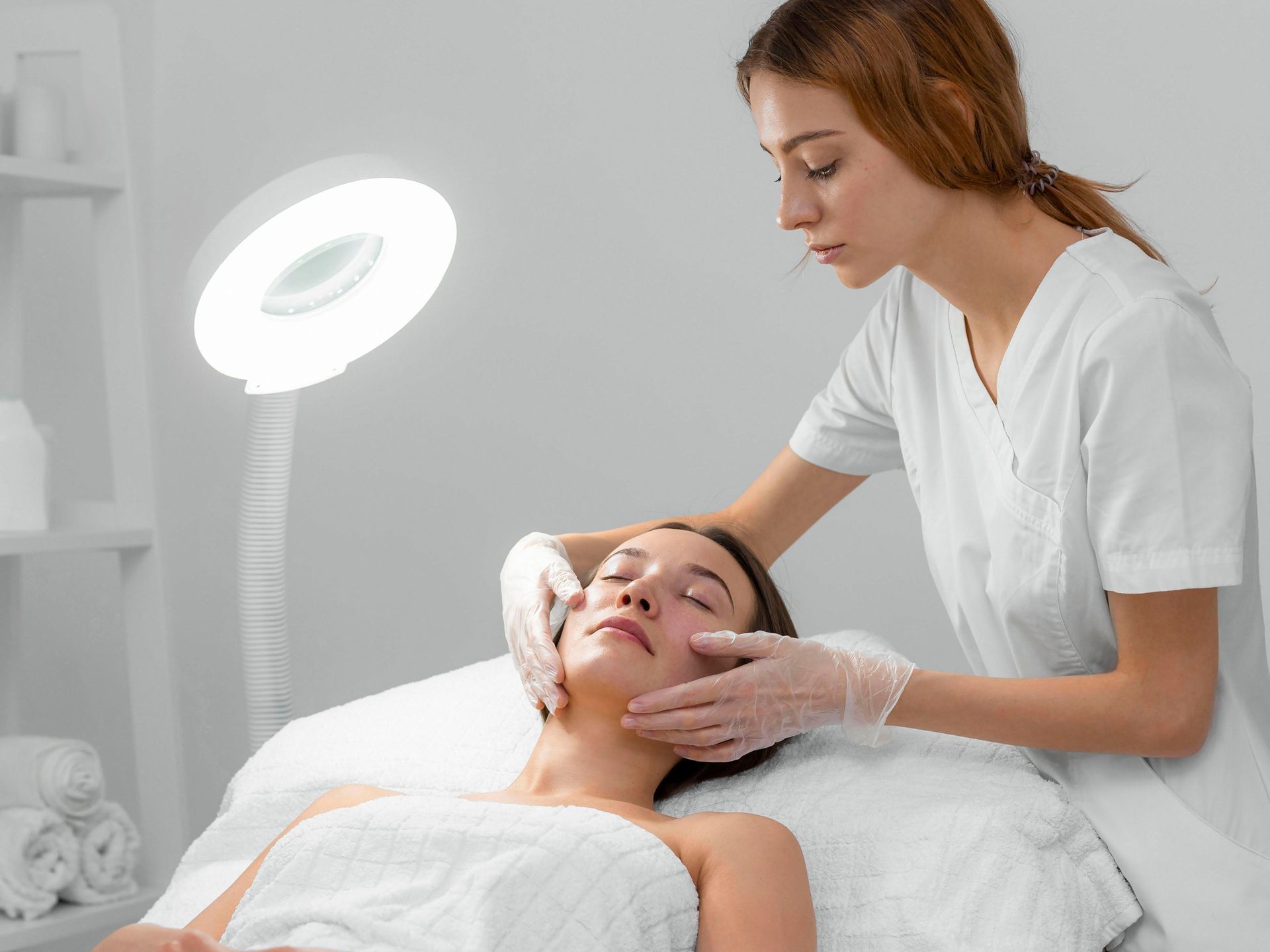 Woman receiving a facial from a professional wearing gloves, under a bright light.