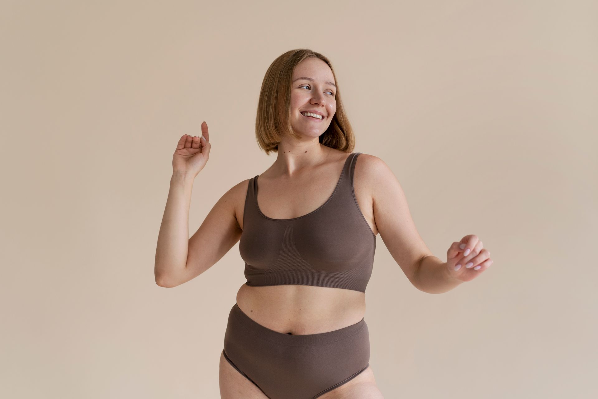 Woman in brown underwear, smiling and dancing against a beige background.