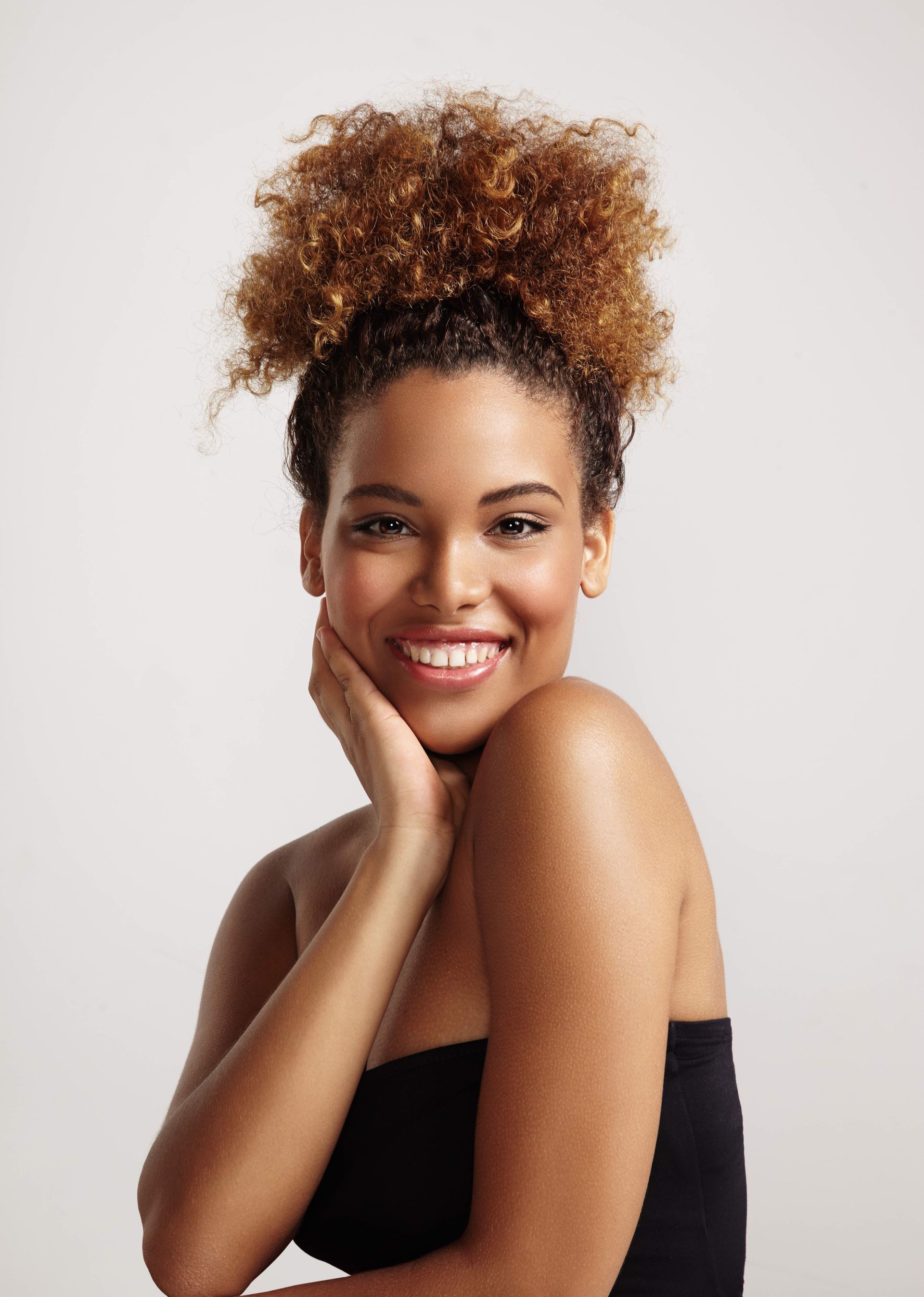 Woman with curly brown hair in a high bun, wearing a black top, smiling with hand on cheek.