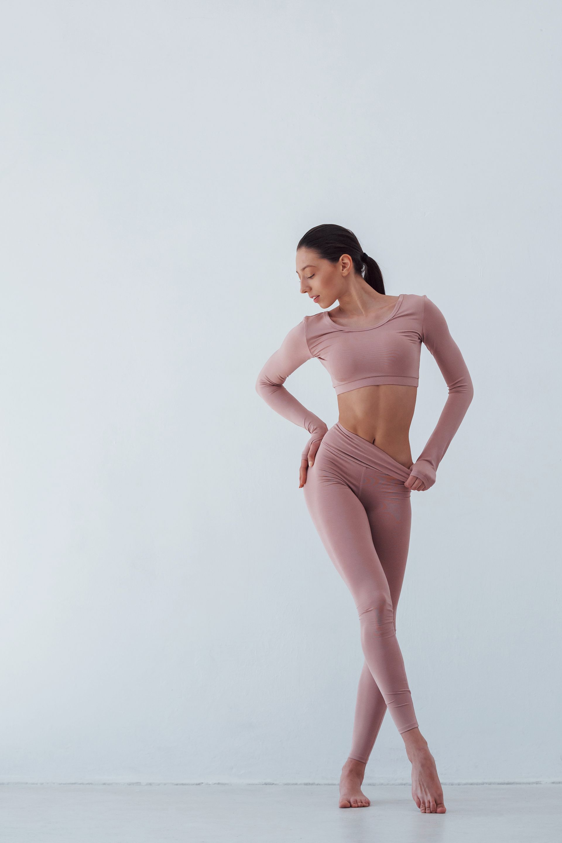 Woman in pink workout clothes, hands on hips, looking down, in front of a white wall.