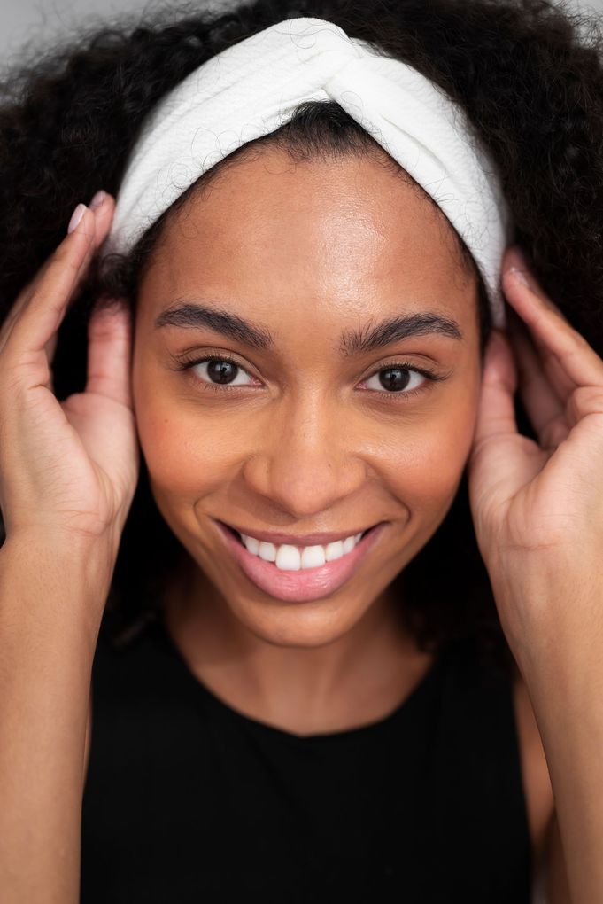 Woman smiling, wearing white headband, holding sides of face, dark hair, black top.