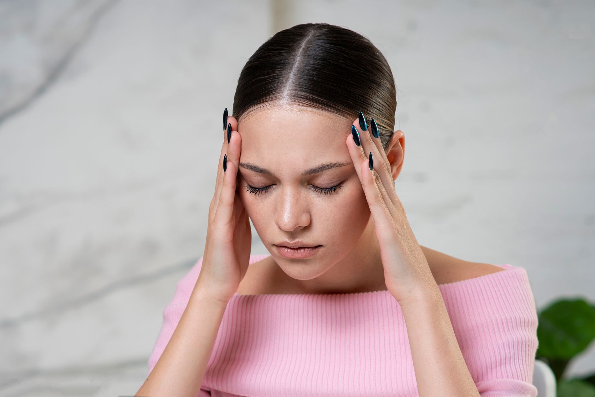 Woman with hands on head, eyes closed, appearing to have a headache, wearing pink top.