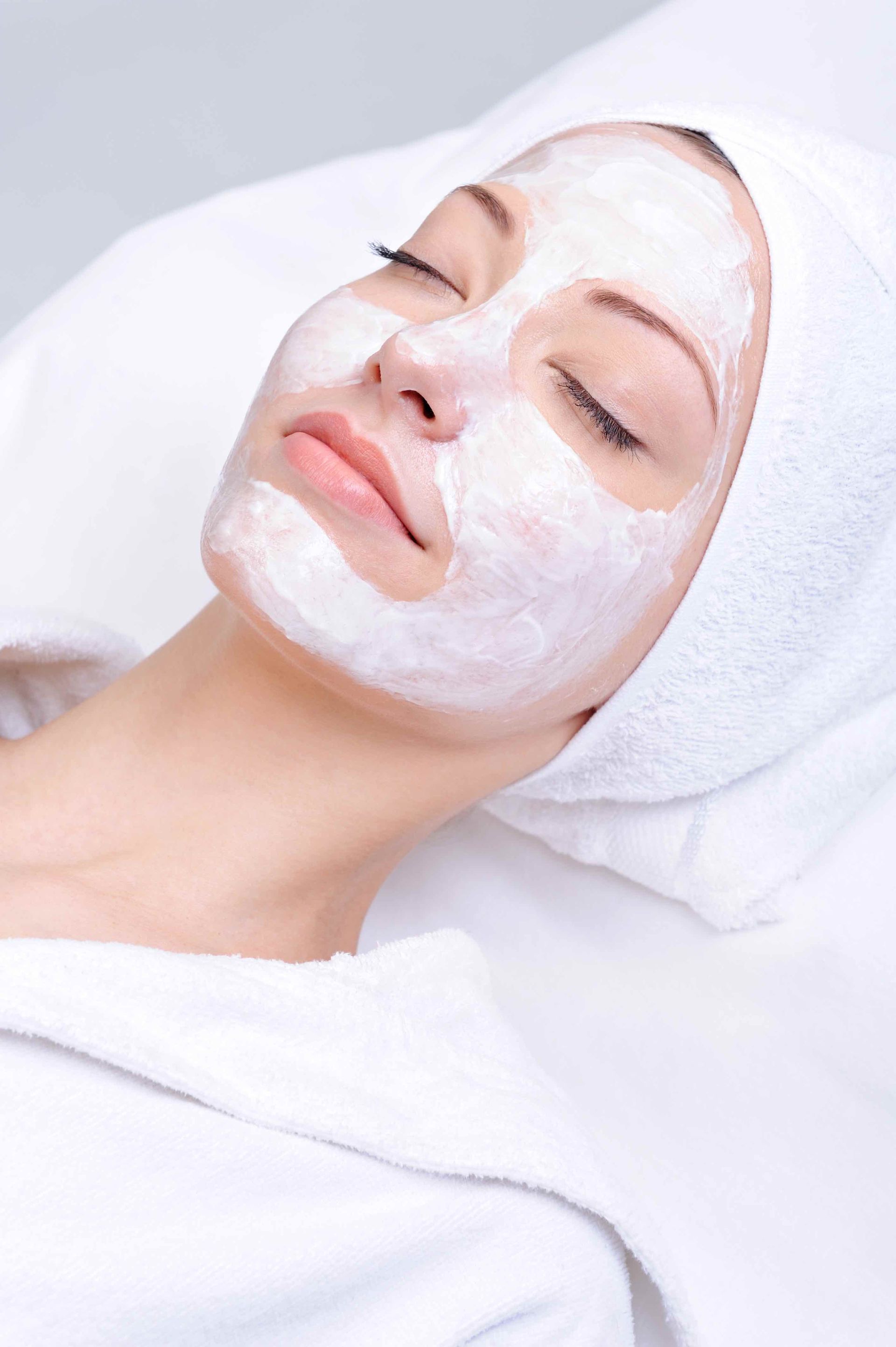 Woman with facial mask, eyes closed, resting on a white towel, wearing a white head wrap.