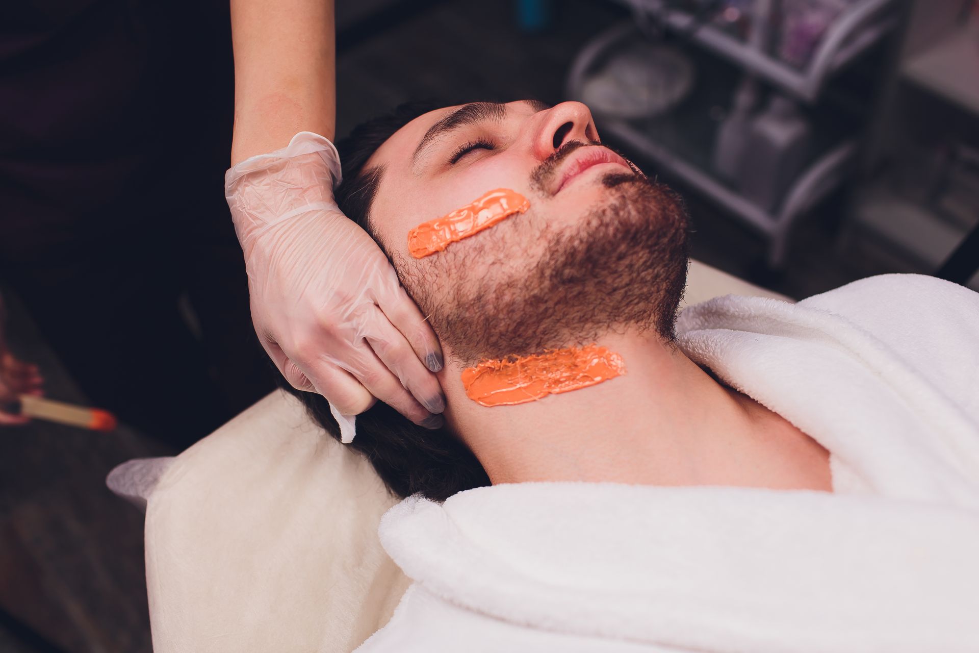 Man receiving facial wax treatment in a salon. Orange wax applied to cheeks and neck.
