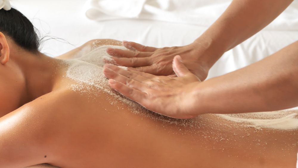 Hands massaging back covered in exfoliating scrub on white spa bed.