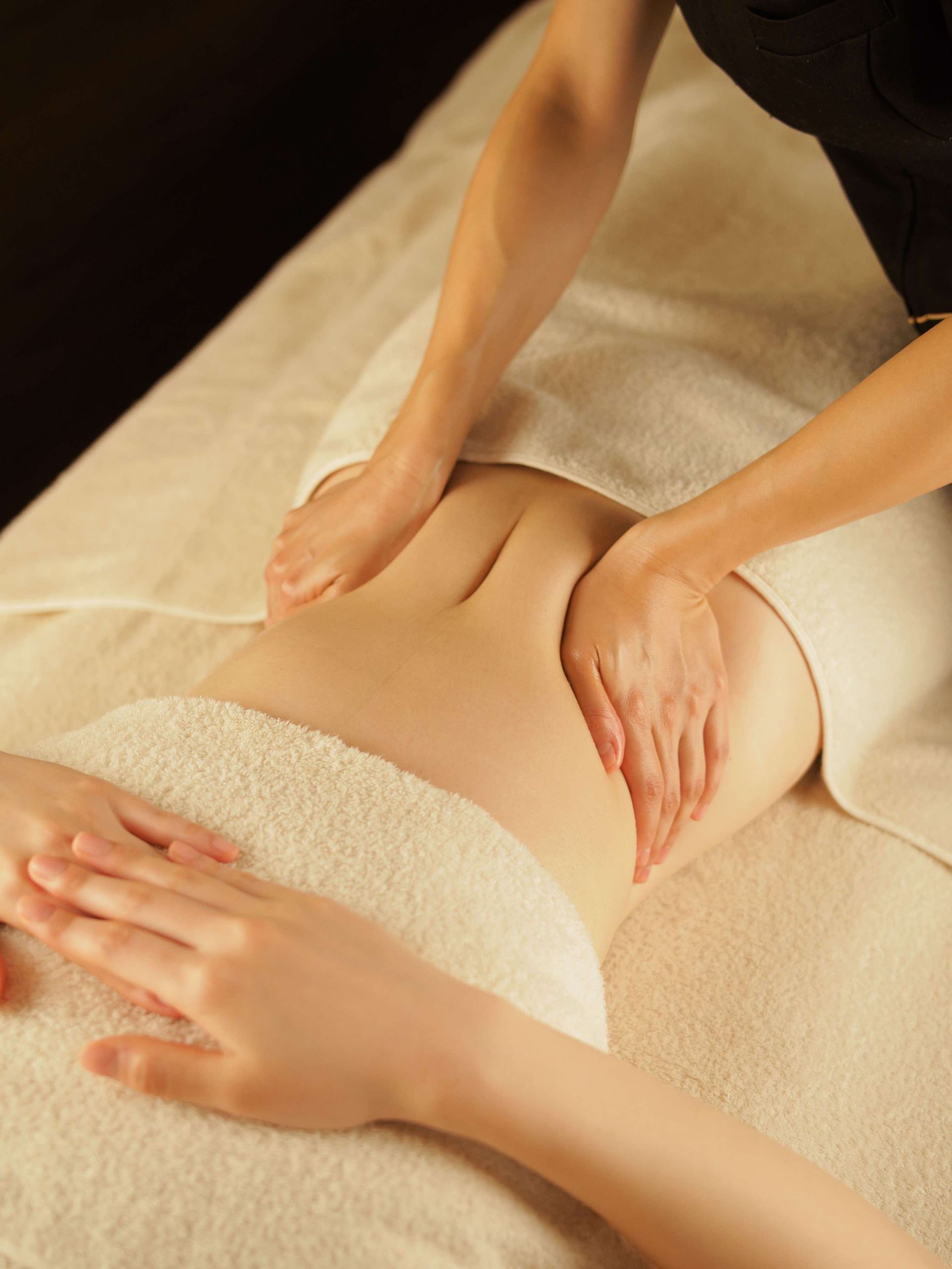 Person receiving abdominal massage, hands on back and stomach, white towels, spa setting.