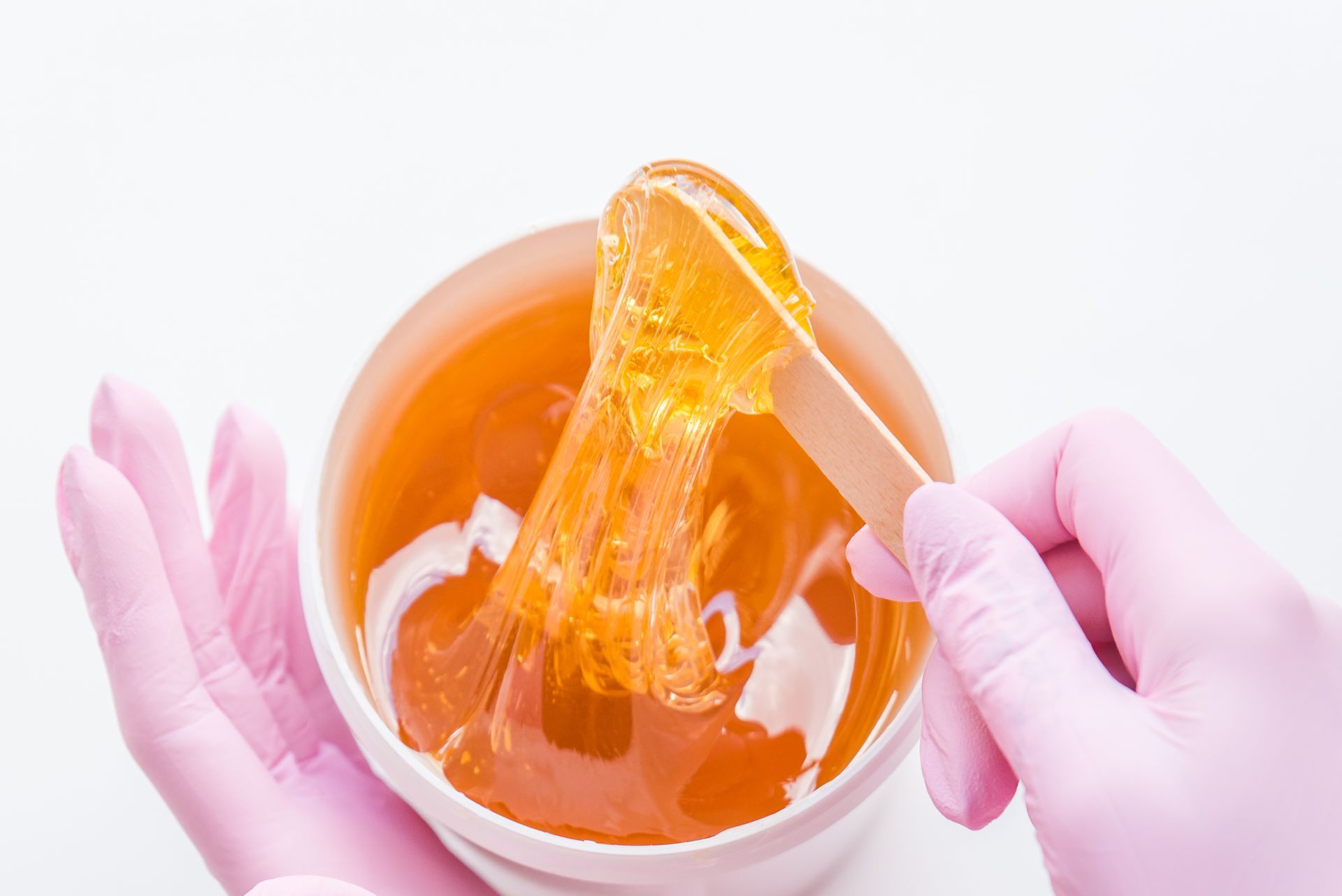 Hands in pink gloves holding a wooden spatula with golden sugar wax over a white container.