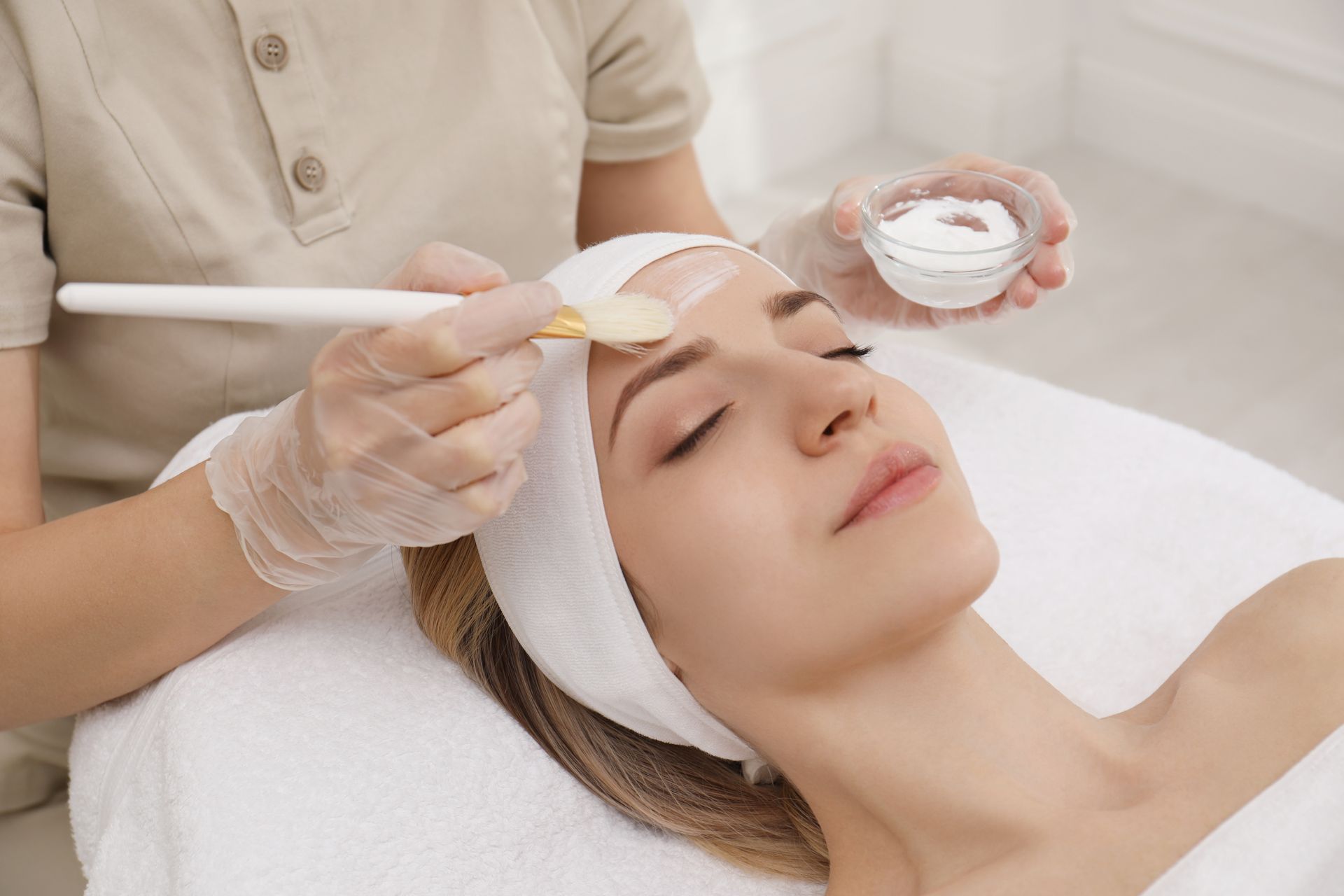 Facial mask being applied at spa. Person lies on a white towel with a headband, receiving treatment.
