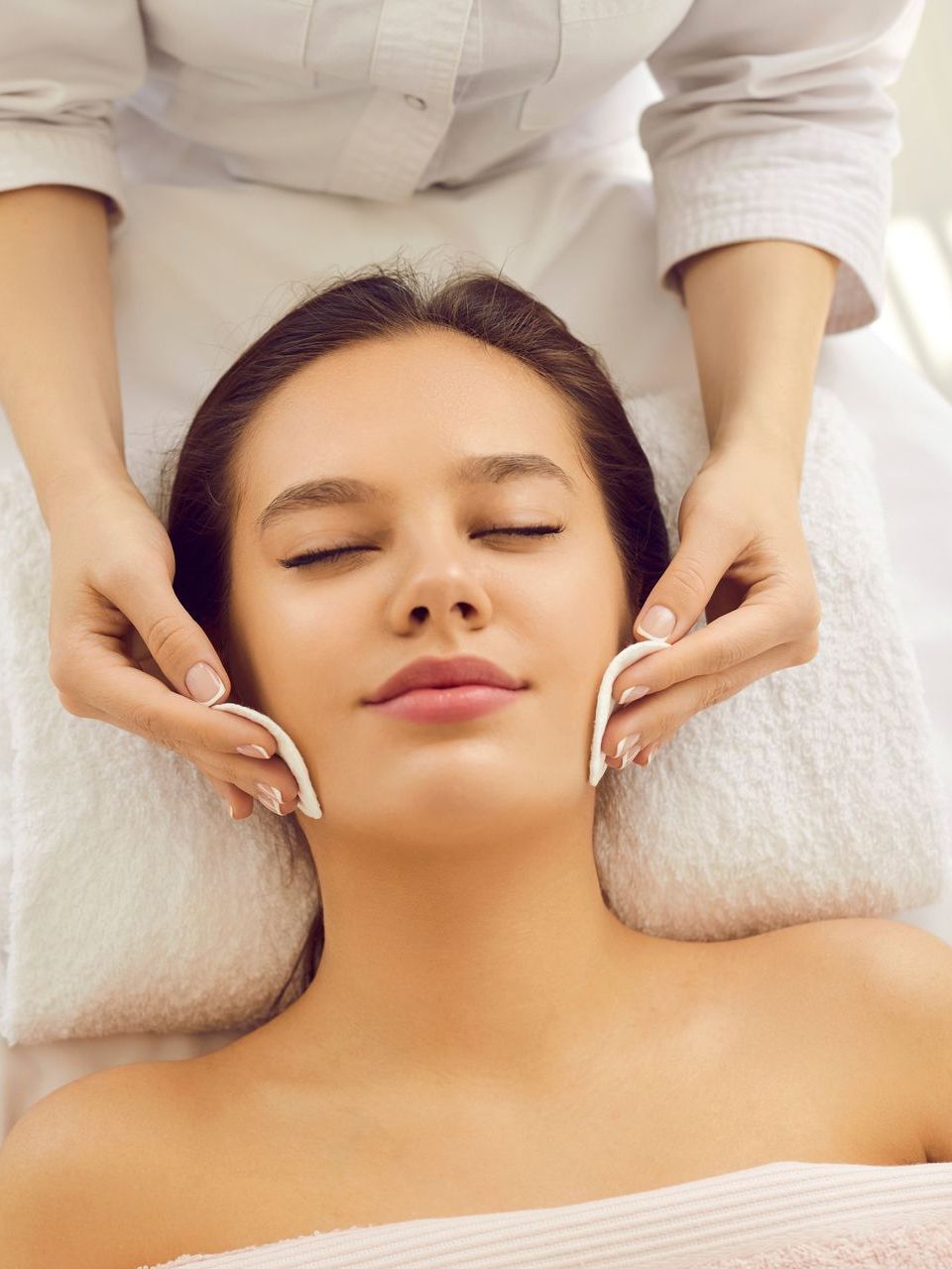 Woman receiving facial treatment, with esthetician applying cotton pads.