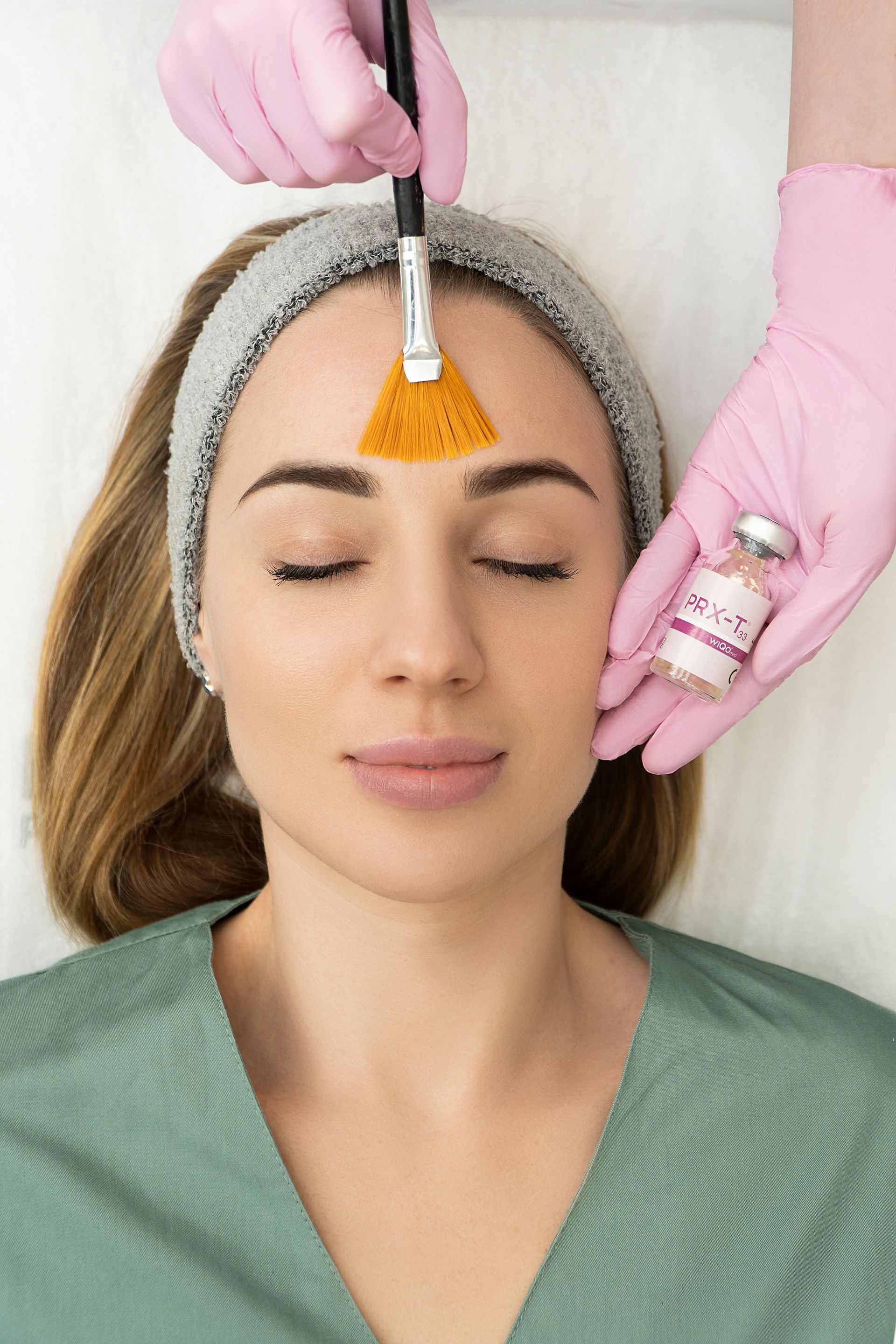 Woman receiving a facial peel, with a brush applying a chemical solution to her forehead, pink-gloved hands, and small vial.