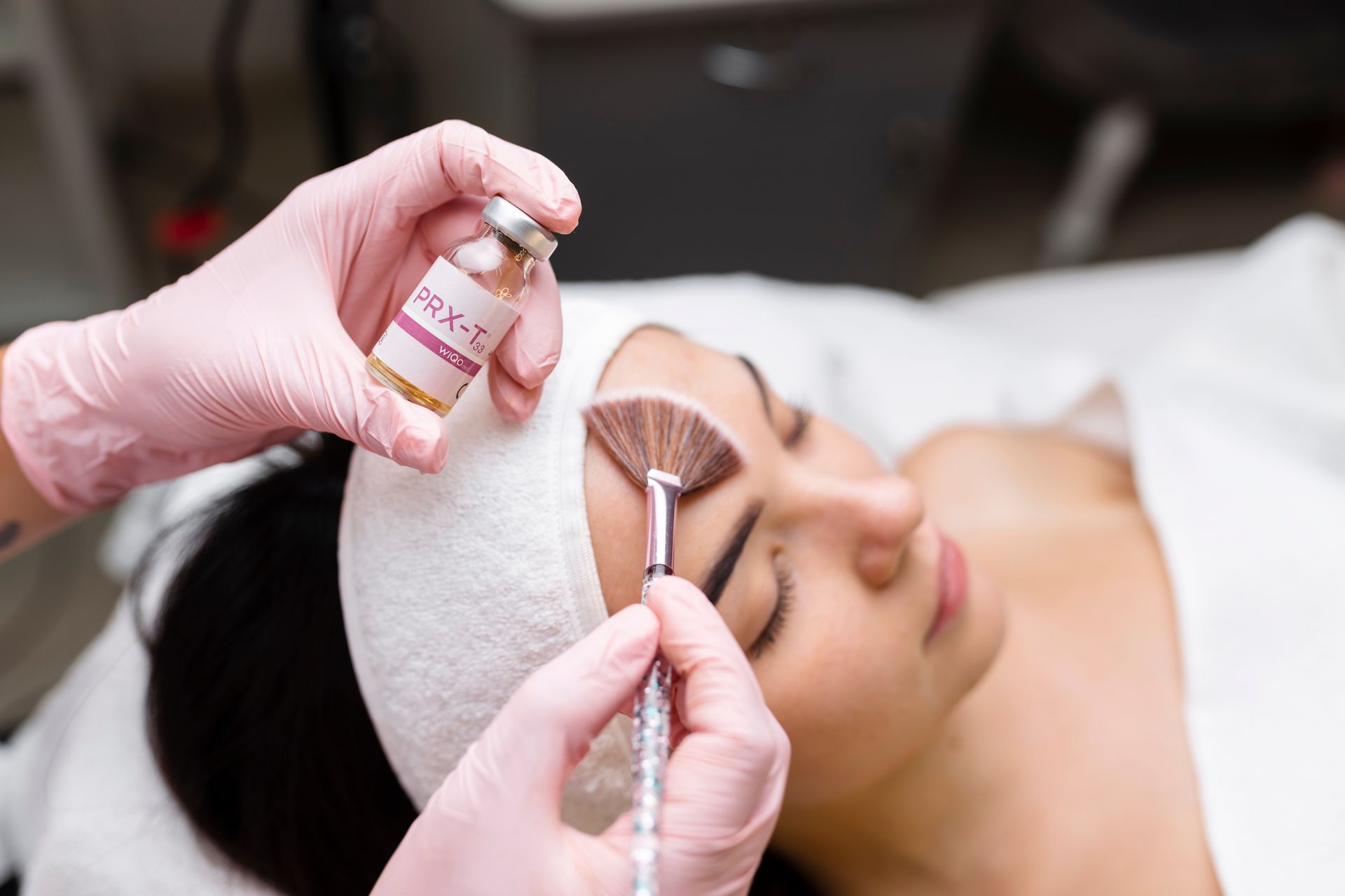 Person receiving a facial treatment; gloved hand applying product with a brush.