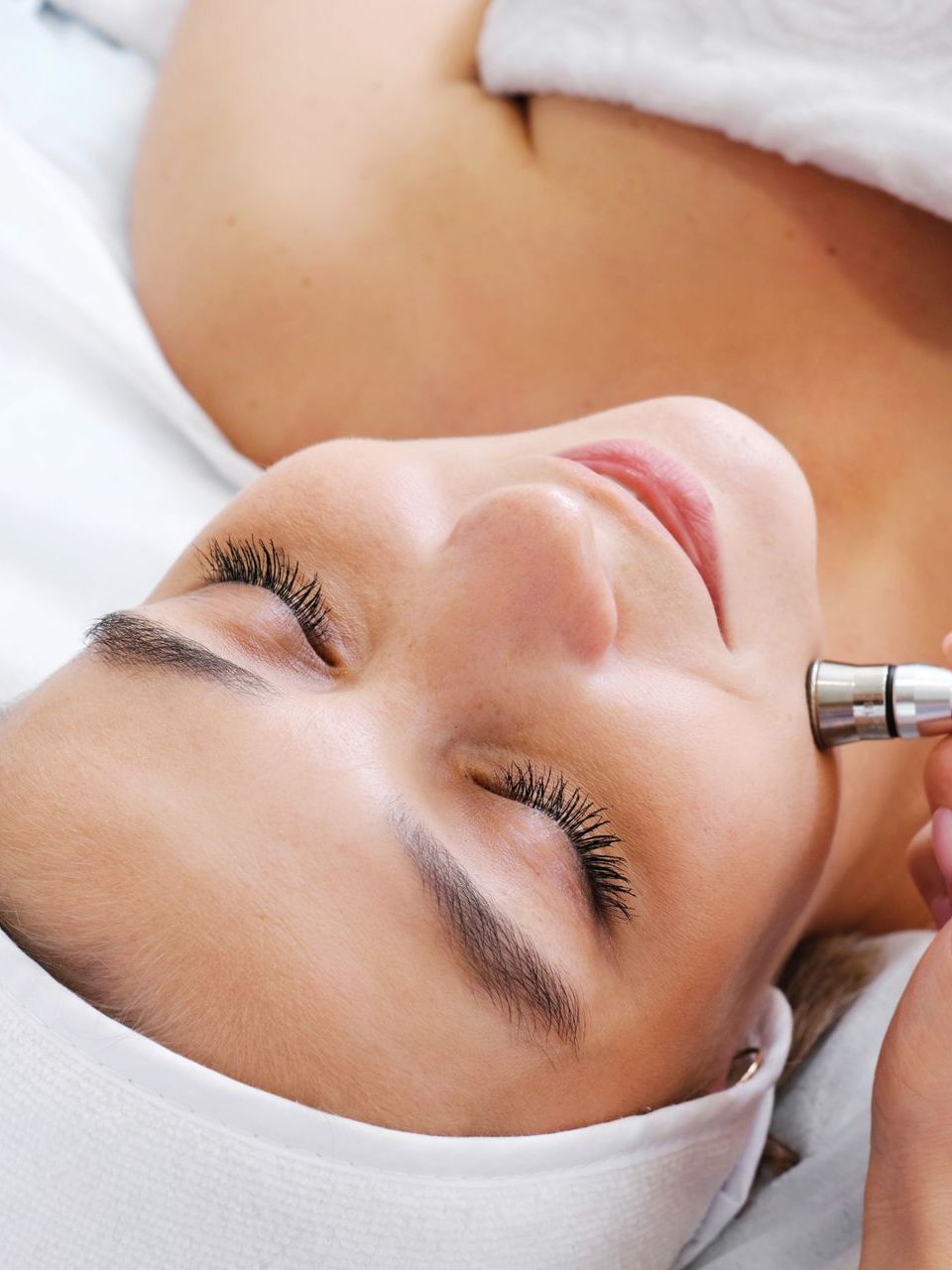 Woman receiving facial treatment; tool on cheek, white cloth, eyes closed.