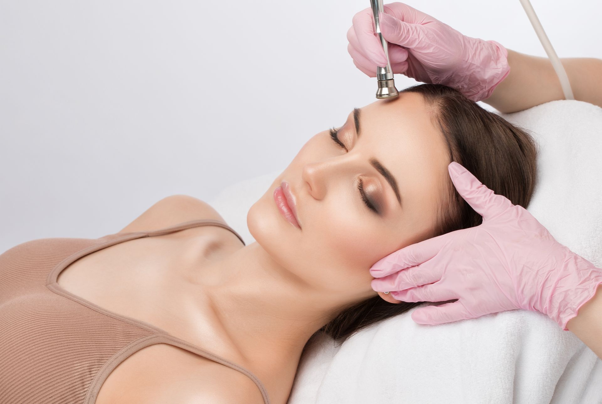 Woman receiving facial treatment; cosmetician with gloved hands using a tool on her forehead.