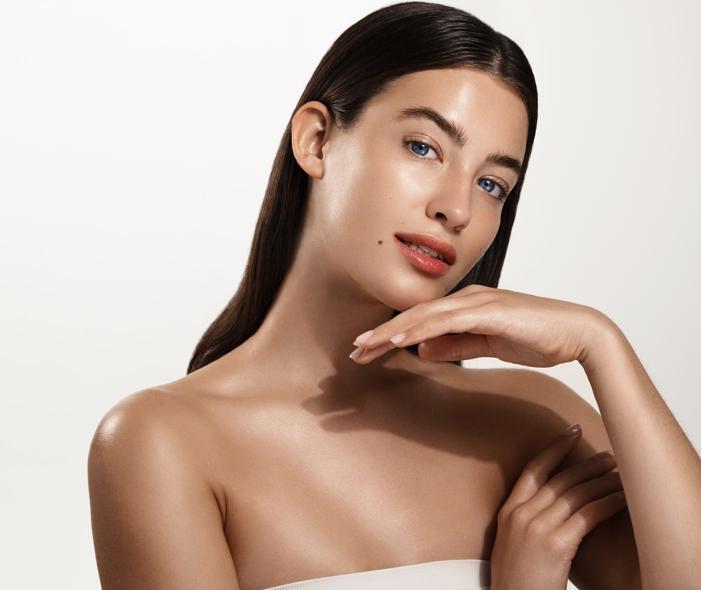 Woman with smooth, glowing skin, long dark hair, lightly tanned. She touches her neck and looks toward the viewer.