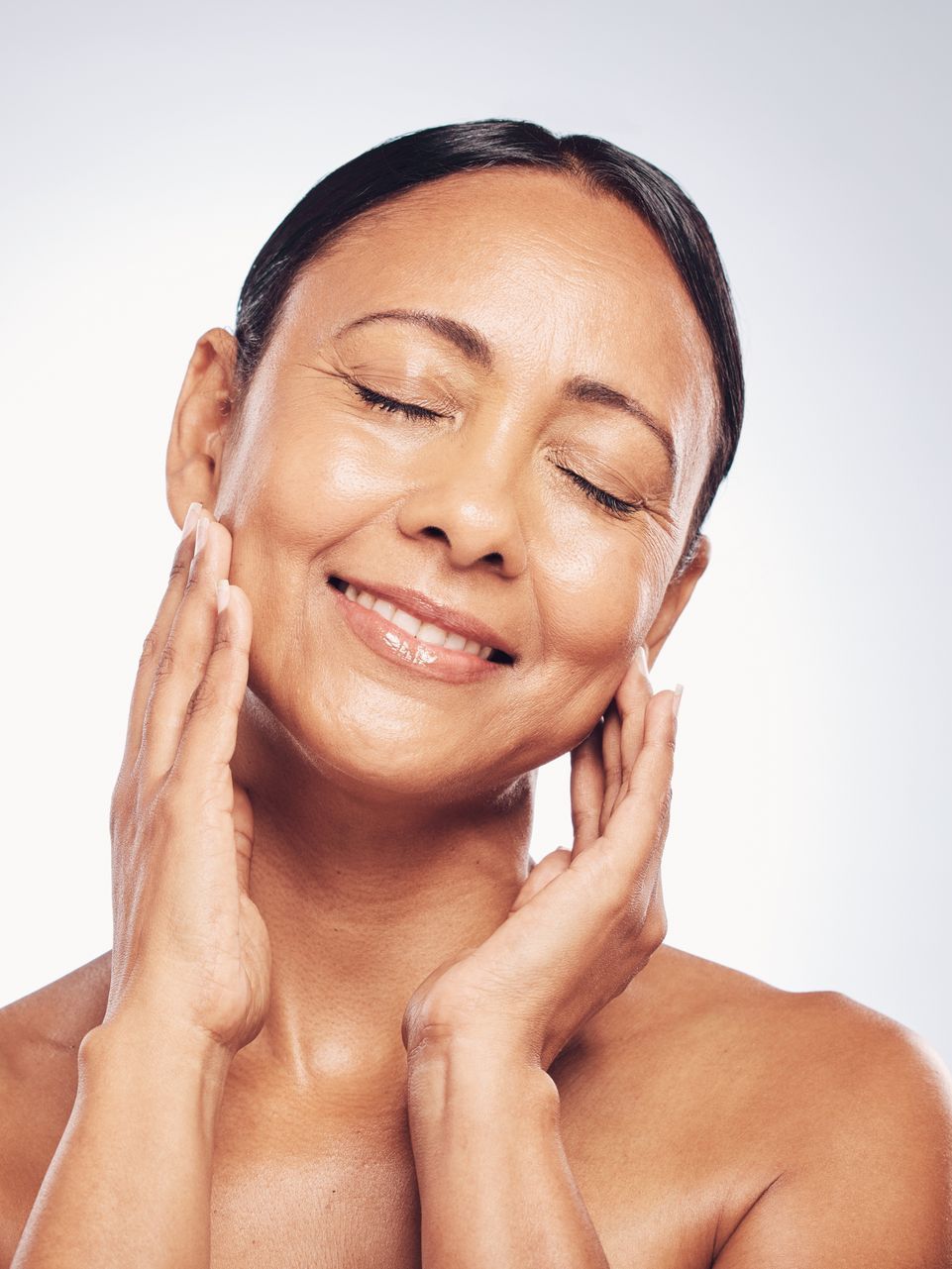 Woman with closed eyes smiling, touching her face with both hands, soft skin, neutral background.
