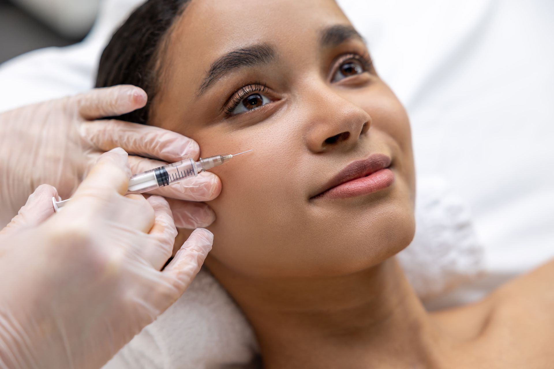 A person receiving an injection in their cheek while lying down. Gloves, syringe, and light skin tones are visible.