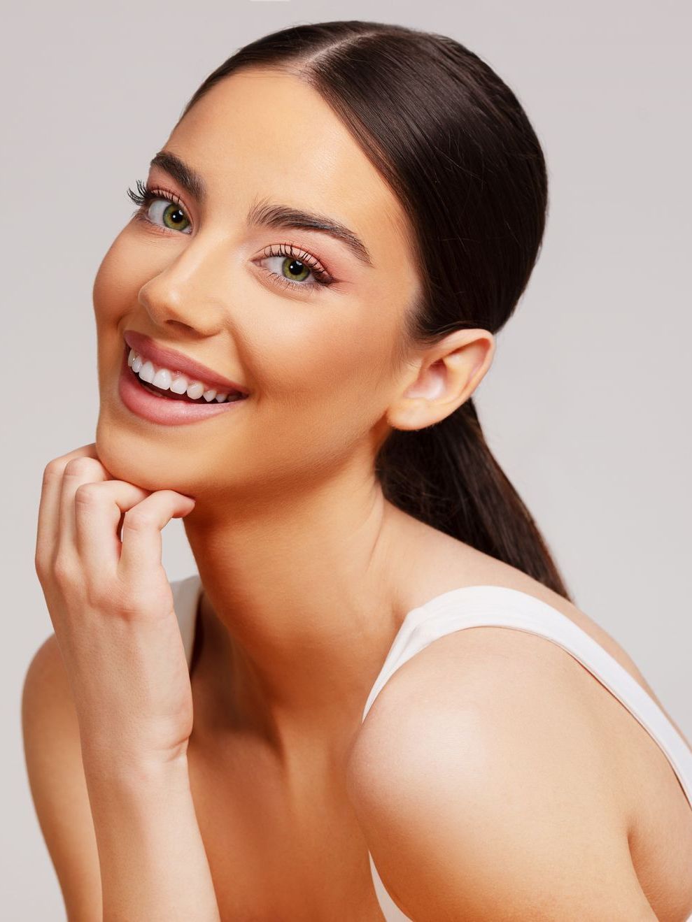 Woman smiles, hand on chin, wearing a white top. Natural makeup, dark hair pulled back.