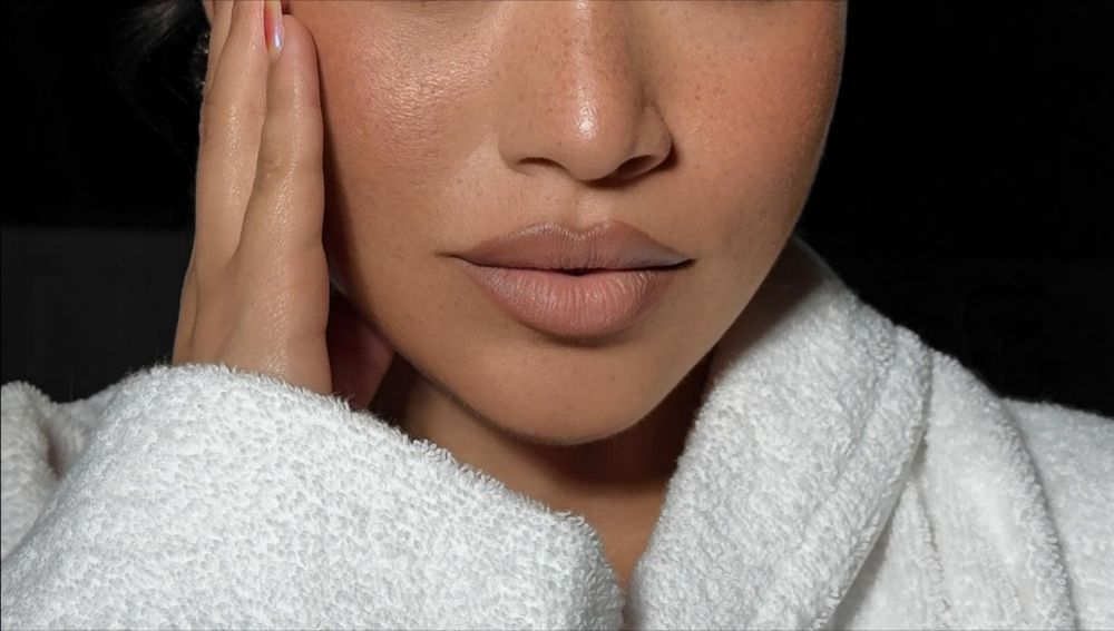 Woman's face with nude lipstick, resting hand on cheek. Wearing white robe. Soft lighting.