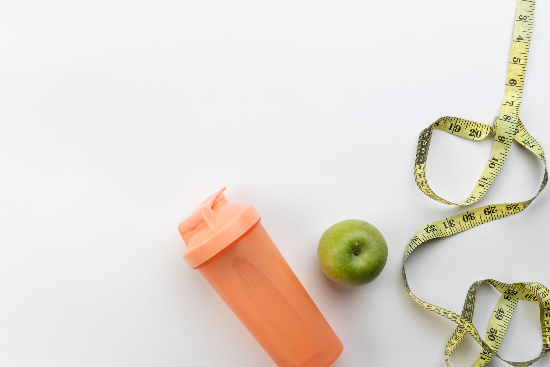 Orange protein shaker, green apple, and yellow measuring tape on white surface.
