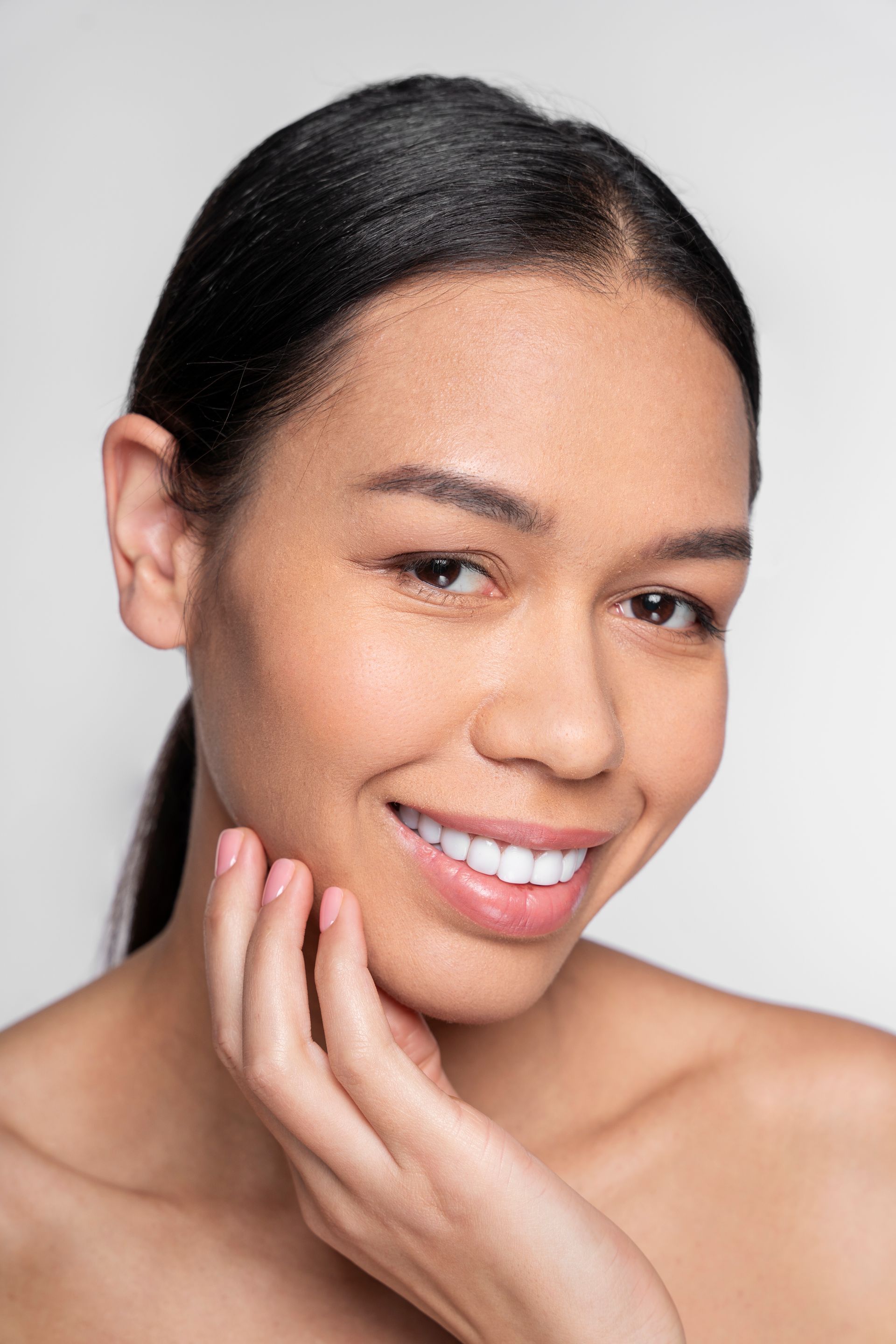 Woman smiling, hand near face, bare shoulders, black hair pulled back, against white background.