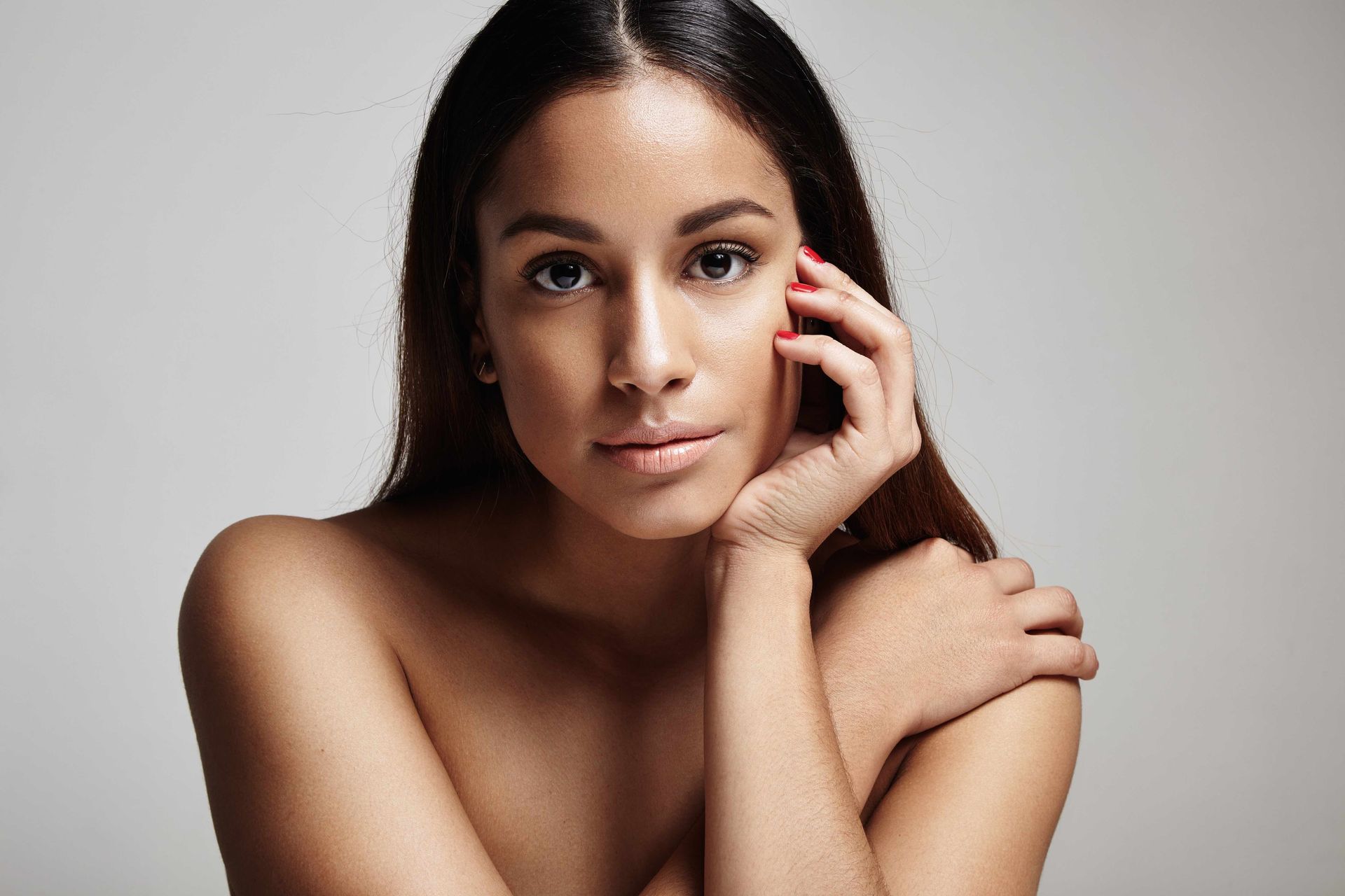 Woman with brown skin, hand on cheek, looking at the camera. Neutral background.