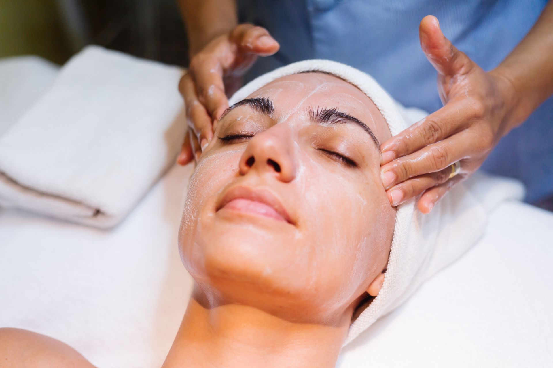 Woman receiving facial treatment, eyes closed, with product on face, hands massaging cheeks.